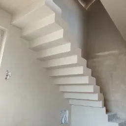 Wykończone, wiszące schody betonowe z tynkowanymi ścianami, widoczne niedoskonałości i kable wystające ze ściany.