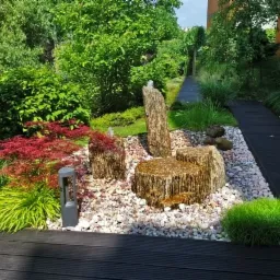 Ogród z minimalistyczną fontanną kaskadową z kamienia, otoczoną białym żwirem, czerwonym klonem palmowym i czarnym tarasem kompozytowym.