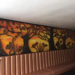 Duży mural przedstawiający drzewa na tle zachodzącego słońca, wykonany w odcieniach pomarańczy i brązu, umieszczony na ścianie nad brązową, pikowaną kanapą w lokalu.