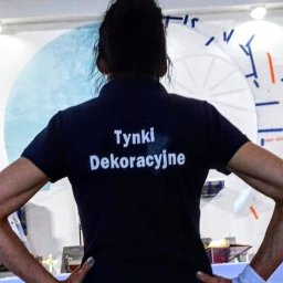 Marta Karpisz - tynkowanie dekoracyjne - Osoba w czarnej koszulce polo z napisem 'Tynki Dekoracyjne' na plecach, stojąca na tle ściany z nieukończonym wzorem koła.