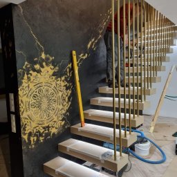 Marta Karpisz - tynkowanie dekoracyjne - Montaż schodów z podświetleniem LED i złotymi balustradami na tle ściany z dekoracyjnym, ciemnym tynkiem i złotym ornamentem. Widoczny pracownik w trakcie montażu balustrady.