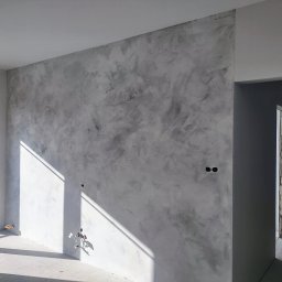 Marta Karpisz - tynkowanie dekoracyjne - Ściana wewnętrzna pokryta dekoracyjnym tynkiem w odcieniach szarości, z widocznymi instalacjami elektrycznymi i fragmentem okna z balkonem w surowym stanie.