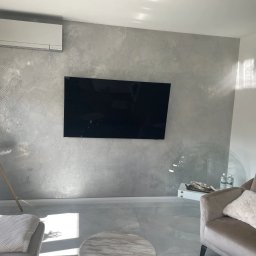 Marta Karpisz - tynkowanie dekoracyjne - Szara ściana z dekoracyjnym tynkiem o wzorze geometrycznym, na której wisi czarny telewizor, klimatyzator pod sufitem, szara podłoga i fragment szarego fotela z poduszką.