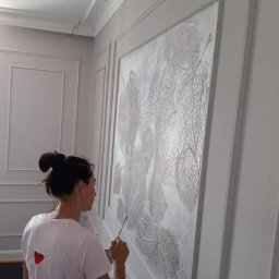 Marta Karpisz - tynkowanie dekoracyjne - Kobieta z upiętymi włosami maluje pędzlem dekoracyjny wzór liści na ścianie z panelami w odcieniach szarości.