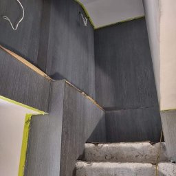 Marta Karpisz - tynkowanie dekoracyjne - Świeżo pomalowana na szaro klatka schodowa z surowymi betonowymi schodami, zabezpieczona taśmą malarską przy suficie i ościeżnicy, widoczne wystające kable elektryczne.
