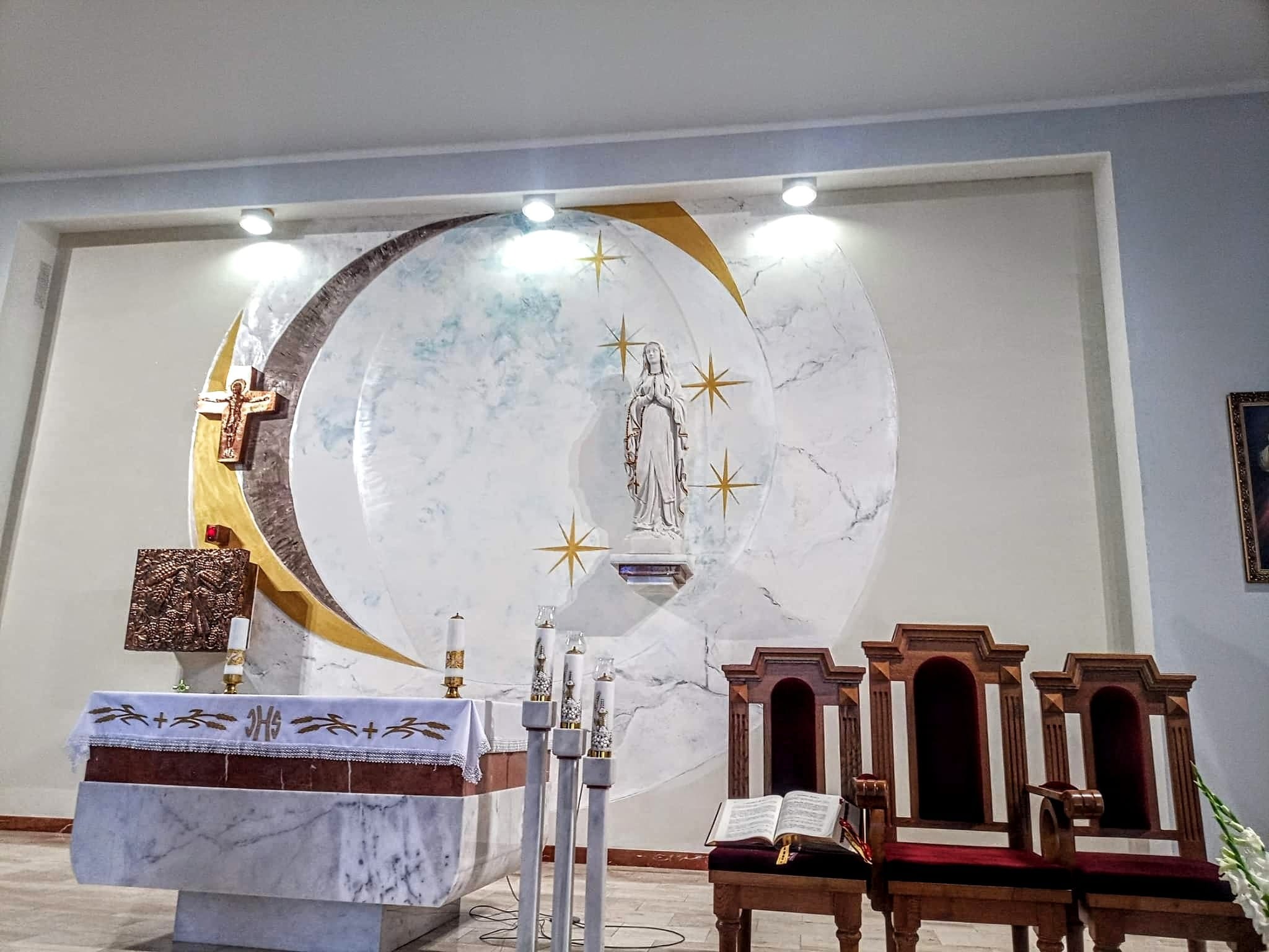 Wnętrze kościoła po renowacji: marmurowy ołtarz z haftowanym obrusem, rzeźba Matki Boskiej na tle dekoracyjnej ściany z motywem księżyca i gwiazd, drewniane krzesła z otwartą księgą.