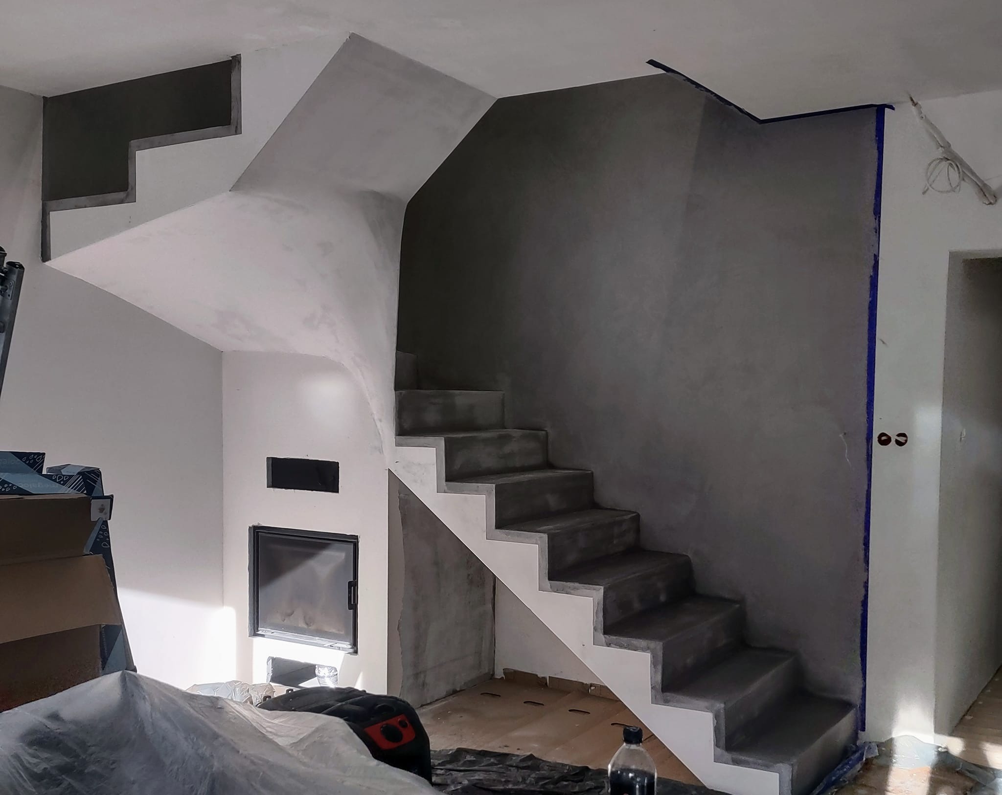 Surowe schody betonowe w trakcie budowy, z widocznymi śladami tynku i taśmą malarską zabezpieczającą ściany, obok kominek z czarną ramą i częściowo przykryte materiałami budowlanymi podłogi.