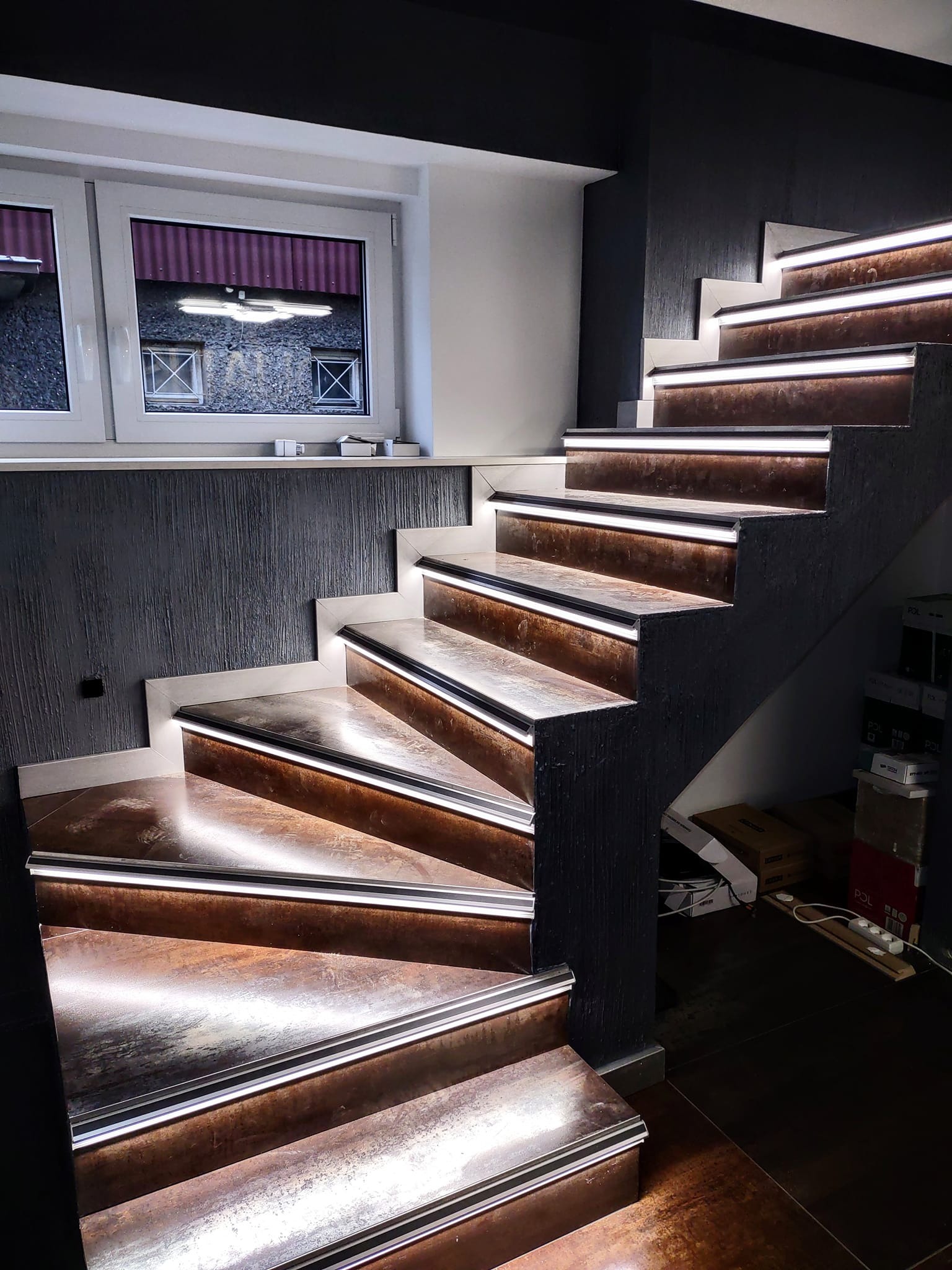 Nowoczesne schody z podświetleniem LED na każdym stopniu, ciemna ściana z fakturą, okno z widokiem na budynek z kratami w oknach.