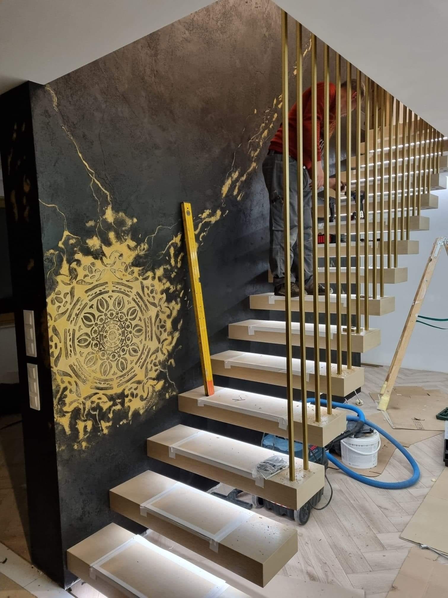 Montaż schodów z podświetleniem LED i złotymi balustradami na tle ściany z dekoracyjnym, ciemnym tynkiem i złotym ornamentem. Widoczny pracownik w trakcie montażu balustrady.