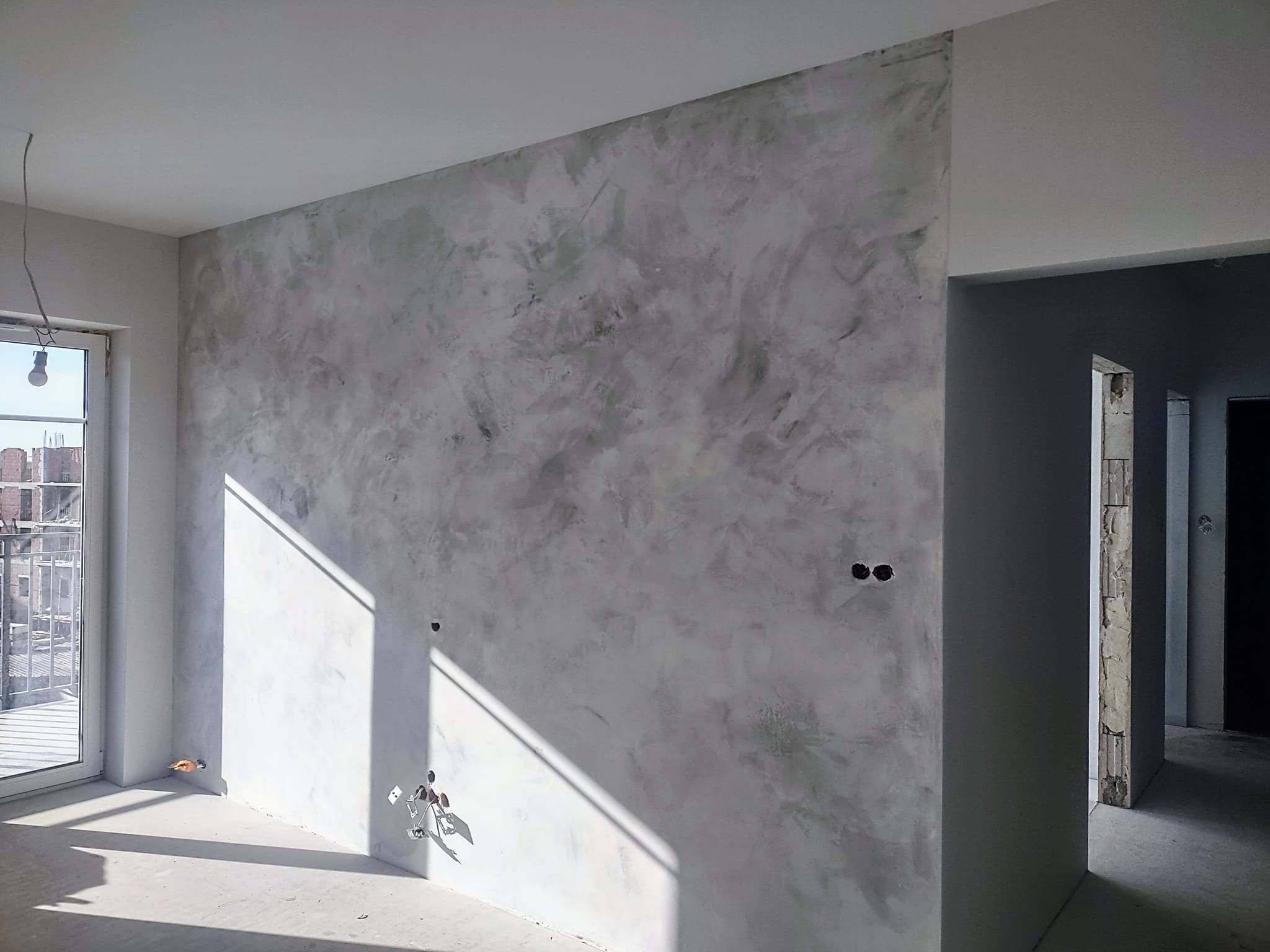 Ściana wewnętrzna pokryta dekoracyjnym tynkiem w odcieniach szarości, z widocznymi instalacjami elektrycznymi i fragmentem okna z balkonem w surowym stanie.