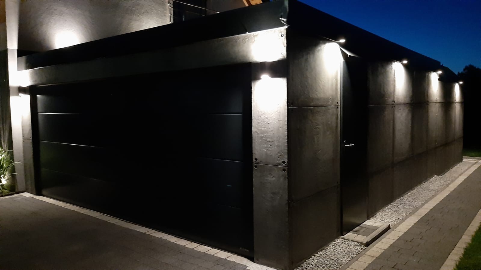 Nowoczesny garaż z czarną bramą segmentową i elewacją z szarych paneli, oświetlony punktowymi lampami zewnętrznymi w nocnym ujęciu.