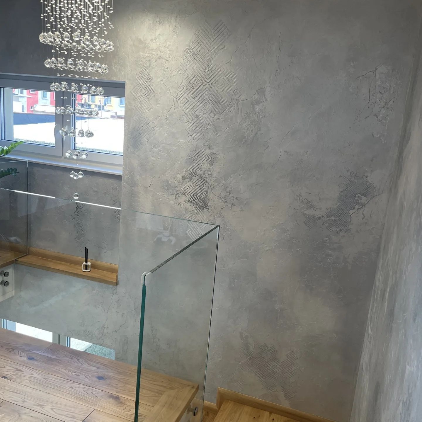 Dekoracyjna ściana w odcieniach szarości z subtelnym geometrycznym wzorem, oświetlona kryształowym żyrandolem, widok z drewnianych schodów z szklaną balustradą.