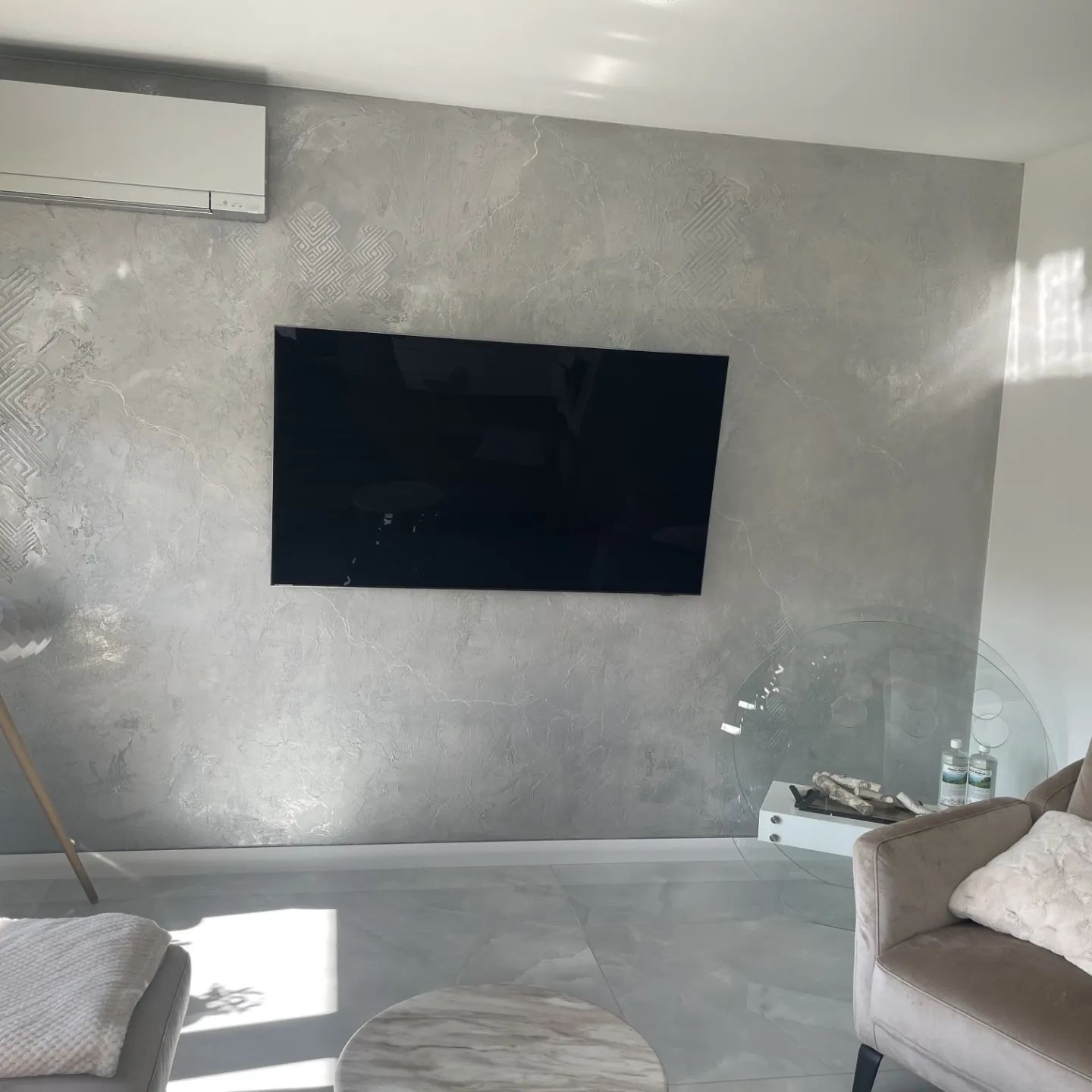 Szara ściana z dekoracyjnym tynkiem o wzorze geometrycznym, na której wisi czarny telewizor, klimatyzator pod sufitem, szara podłoga i fragment szarego fotela z poduszką.