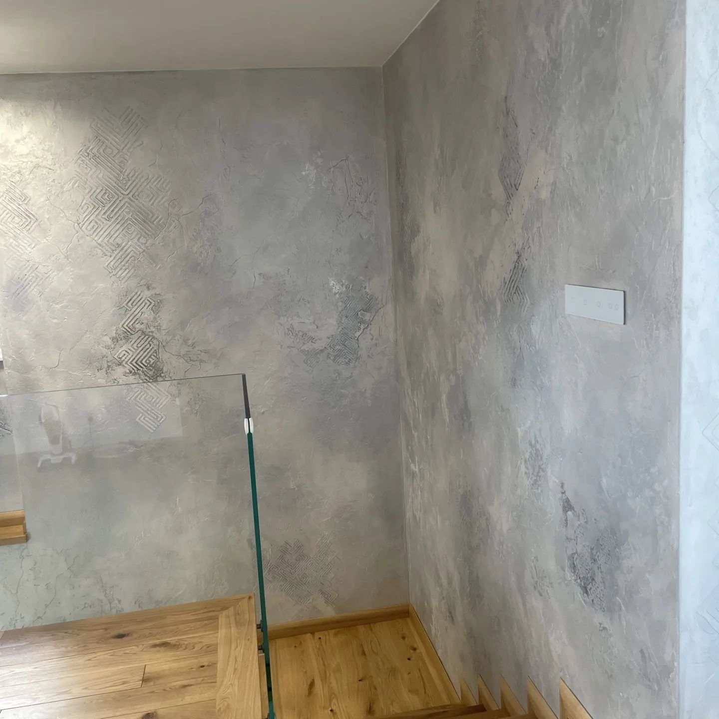 Dekoracyjna ściana w odcieniach szarości z efektem stiuku weneckiego i delikatnym wzorem geometrycznym, widoczna część drewnianych schodów i szklana balustrada.