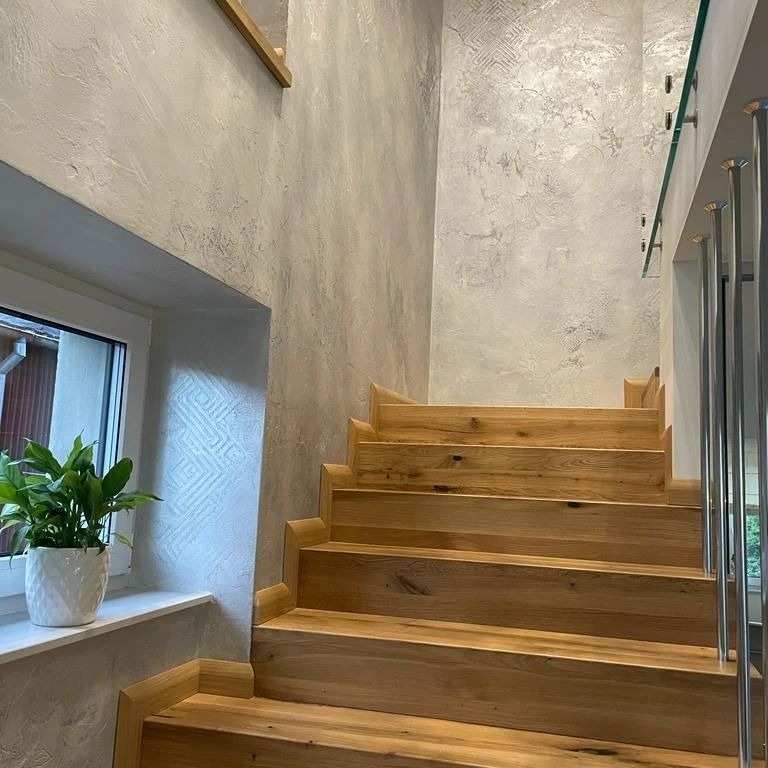 Drewniane schody z ozdobną listwą przypodłogową, prowadzące w górę, z dekoracyjnymi ścianami w odcieniach szarości i rośliną doniczkową w białej osłonce na parapecie okna.