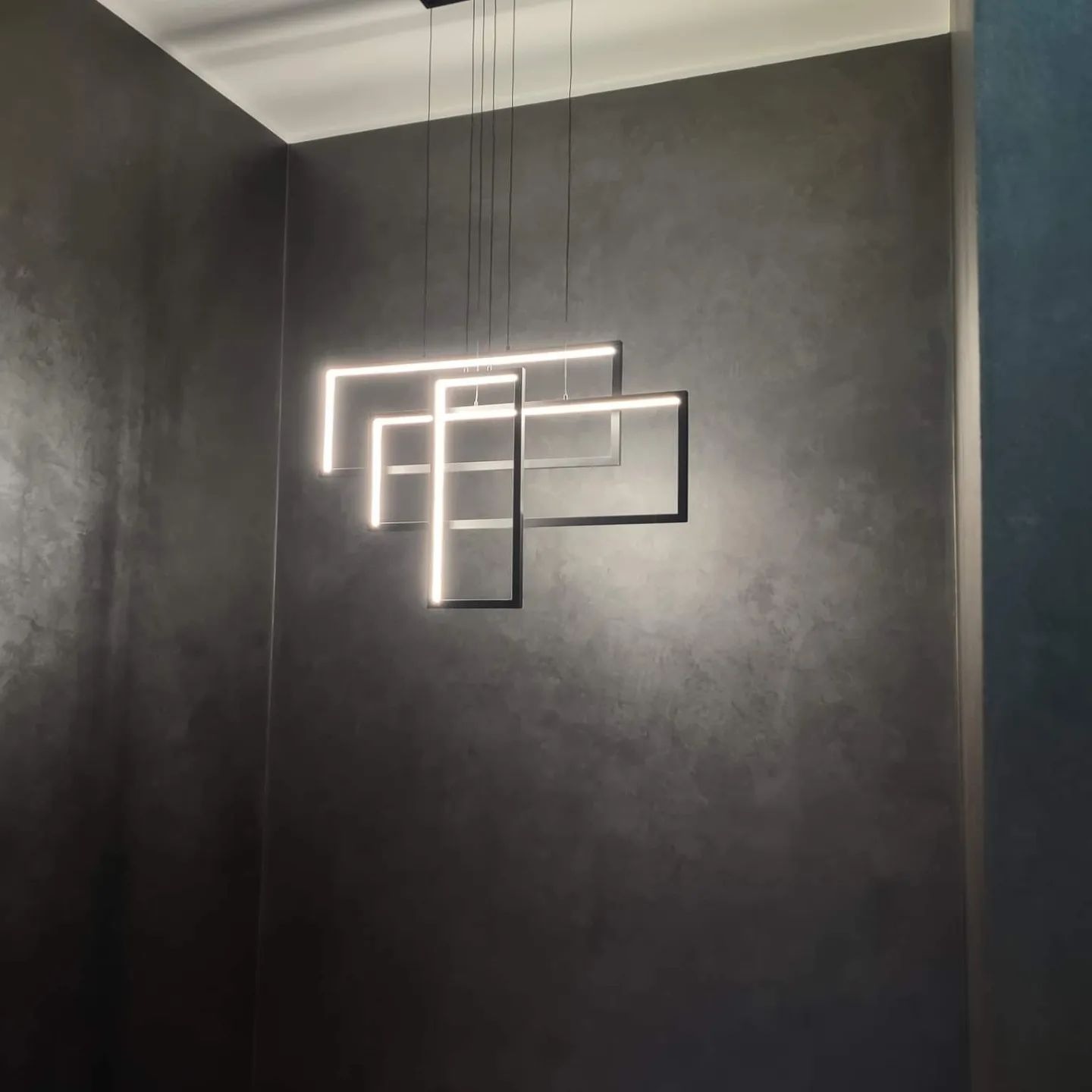 Nowoczesna, geometryczna lampa wisząca z kwadratowymi ramkami LED na tle ściany z efektem betonu architektonicznego w odcieniach szarości.