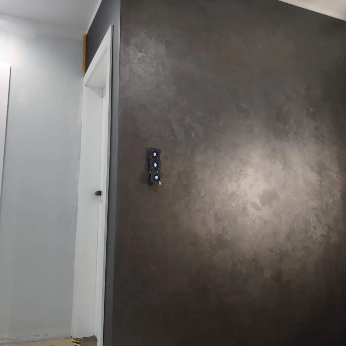 Ściana z dekoracyjną, ciemną fakturą w Zbrosławicach, obok białe drzwi z czarną klamką i szarej ściany. Widoczne zabezpieczone włączniki elektryczne.