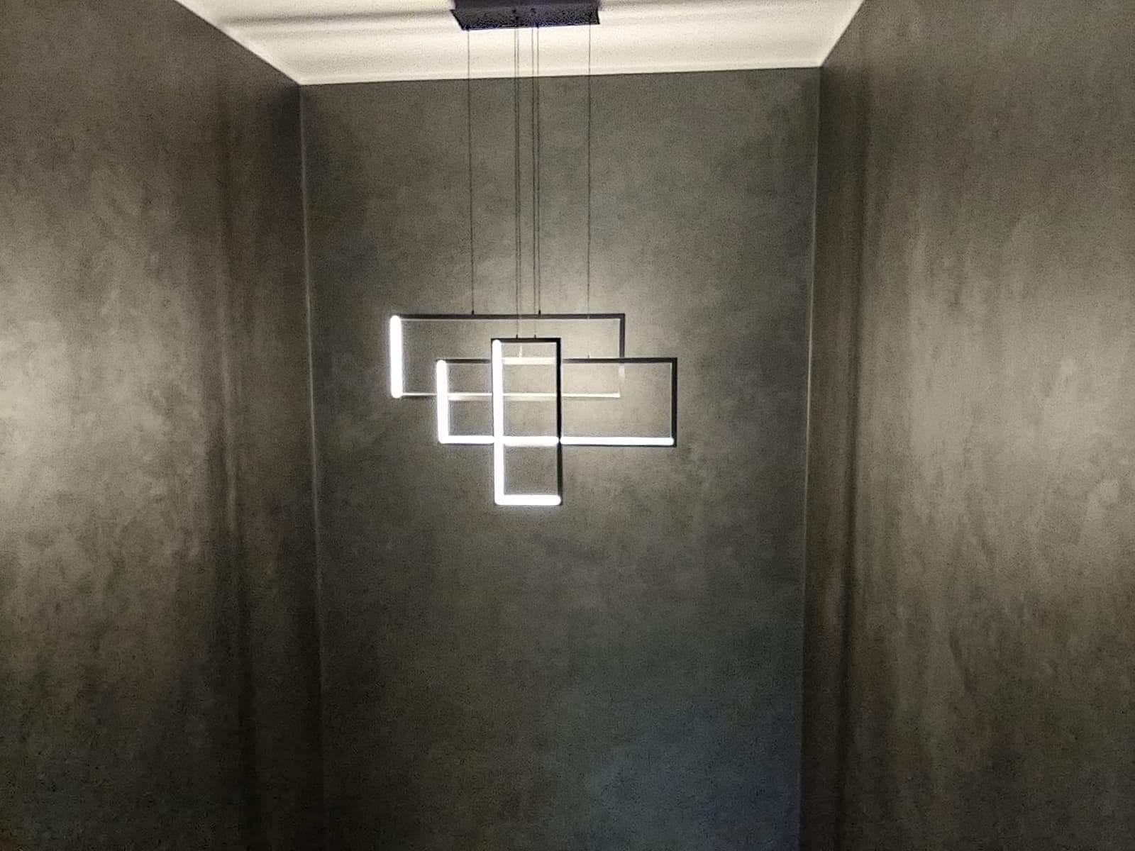 Nowoczesna lampa wisząca z kwadratowych elementów LED na tle ściany o szarej, betonowej fakturze.