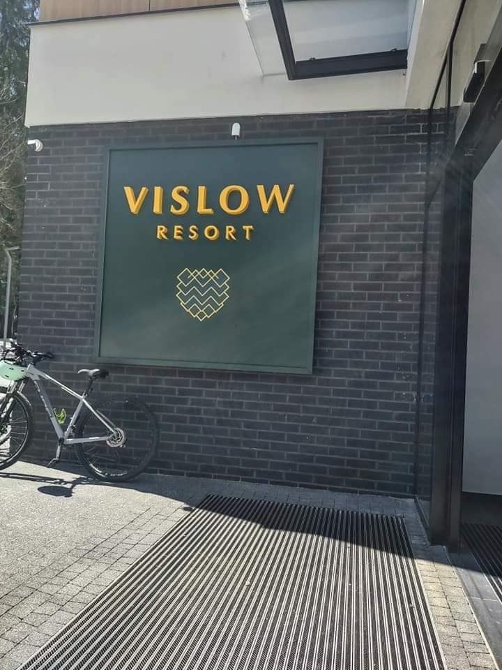 Ciemna ceglana ściana z zielonym szyldem 'VISLOW RESORT' z złotymi literami i geometrycznym logo, rower oparty o ścianę po lewej stronie, kratka odwadniająca przed wejściem.