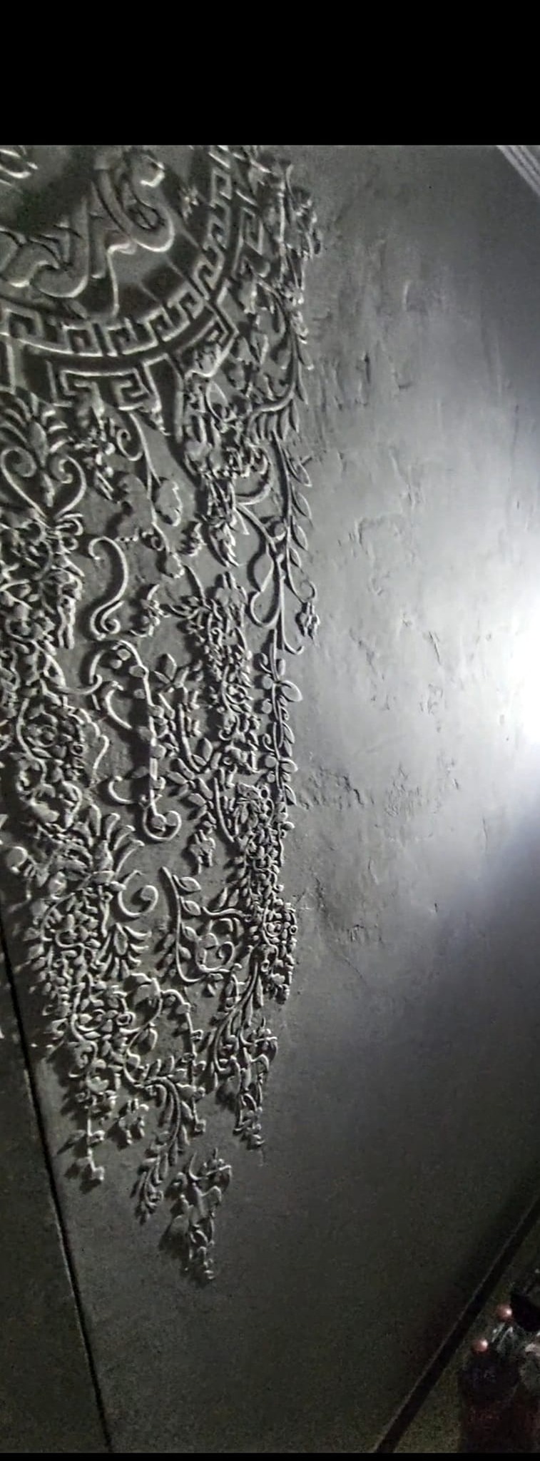 Szara ściana z dekoracyjnym tynkiem strukturalnym przedstawiającym ornament roślinny i geometryczny wzór w górnej części.