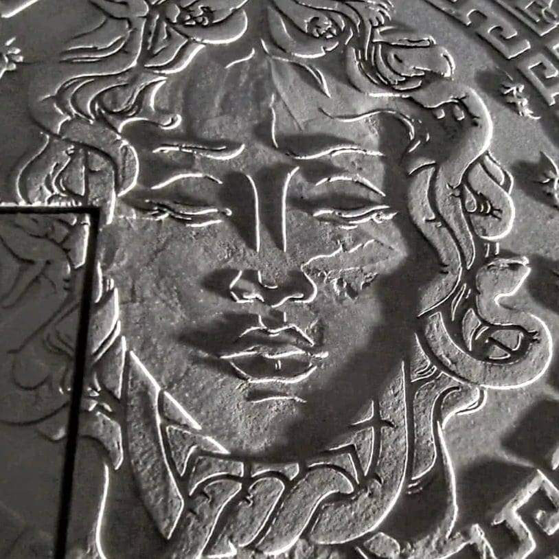 Szara płaskorzeźba przedstawiająca Meduzę z wężami zamiast włosów, oświetlona z góry, tworząca wyraźne cienie.