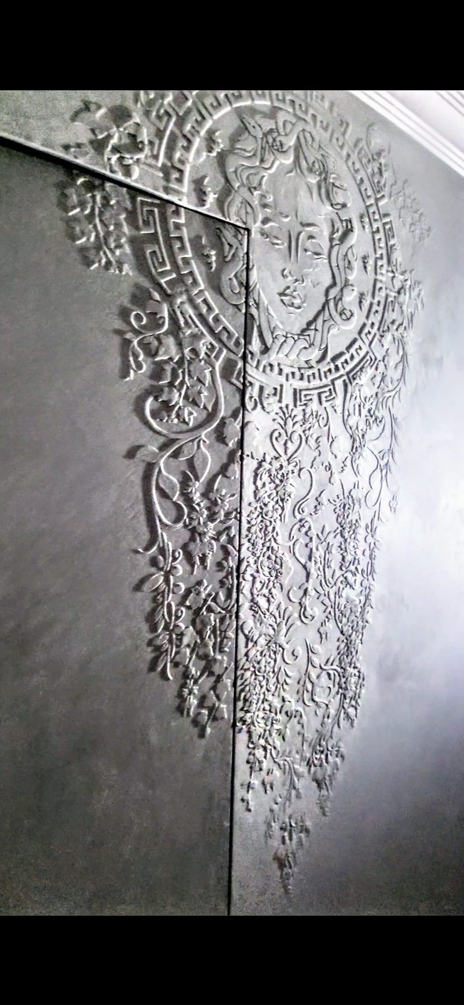 Dekoracyjna ściana z tynkiem strukturalnym przedstawiająca wizerunek Meduzy otoczonej roślinnymi ornamentami, wykonana w odcieniach szarości.