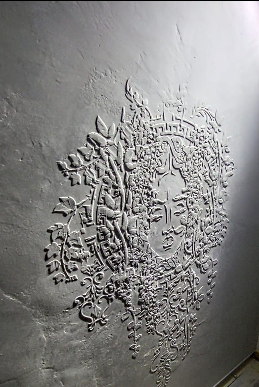 Dekoracyjna płaskorzeźba na szarej ścianie przedstawiająca kobiecą twarz otoczoną ornamentami roślinnymi i geometrycznymi, wykonana w technice tynku strukturalnego.