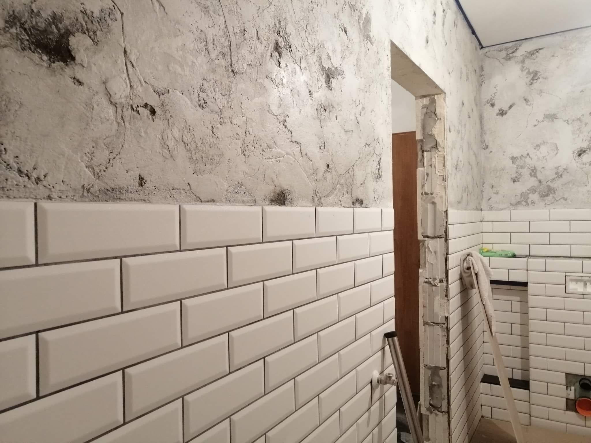 Łazienka w trakcie remontu: białe, prostokątne płytki ułożone do połowy ściany, powyżej surowy tynk imitujący beton, widoczny otwór drzwiowy w stanie surowym, z resztkami obróbki.