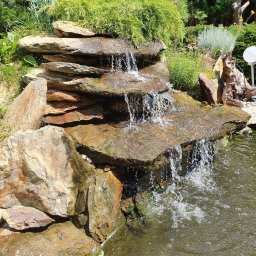 Zielony Folwark - Kaskada wodna z płaskich kamieni w ogrodzie, otoczona zielenią i drzewami, z widocznymi kroplami wody w ruchu.