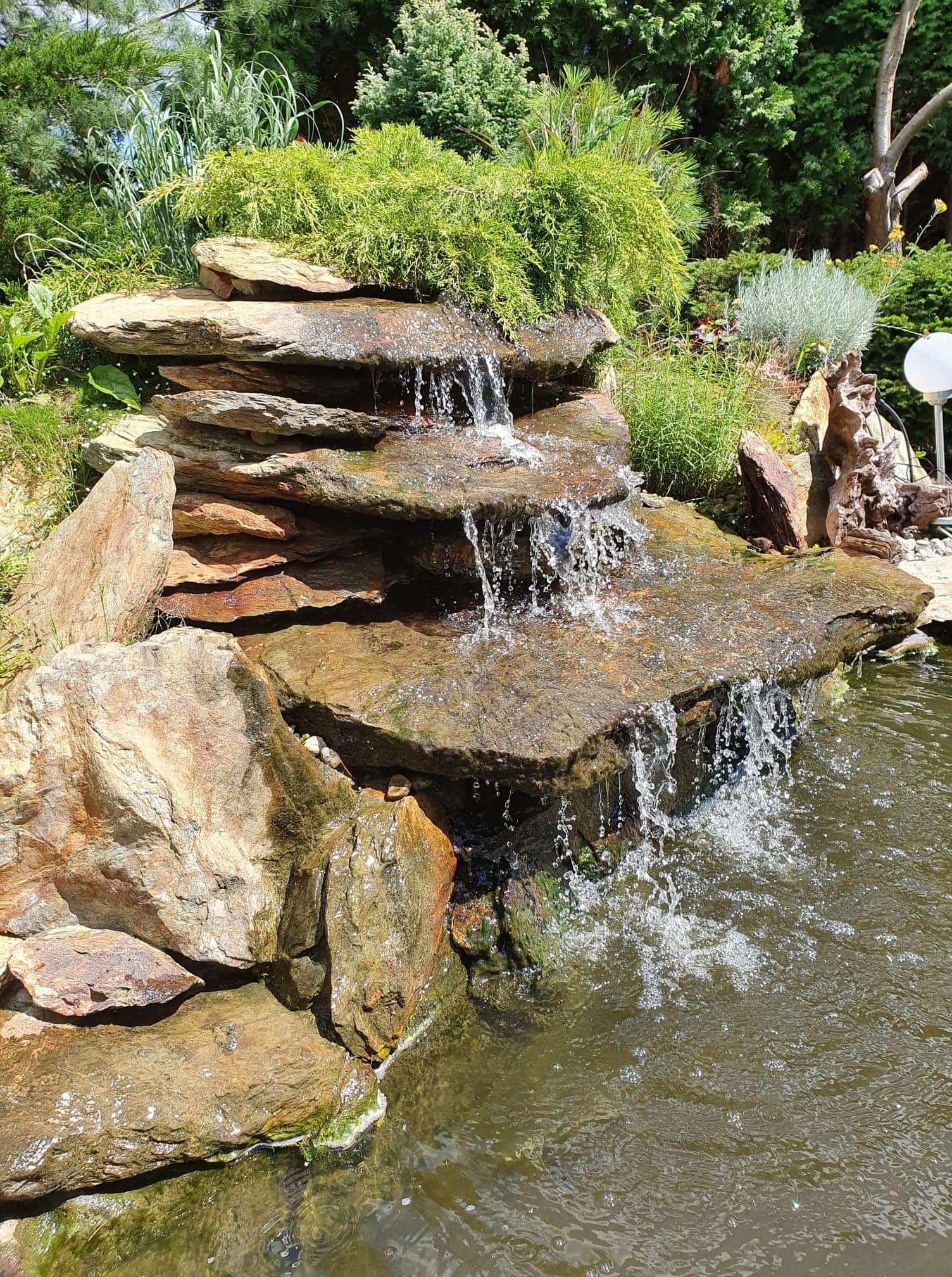 Kaskada wodna z płaskich kamieni w ogrodzie, otoczona zielenią i drzewami, z widocznymi kroplami wody w ruchu.