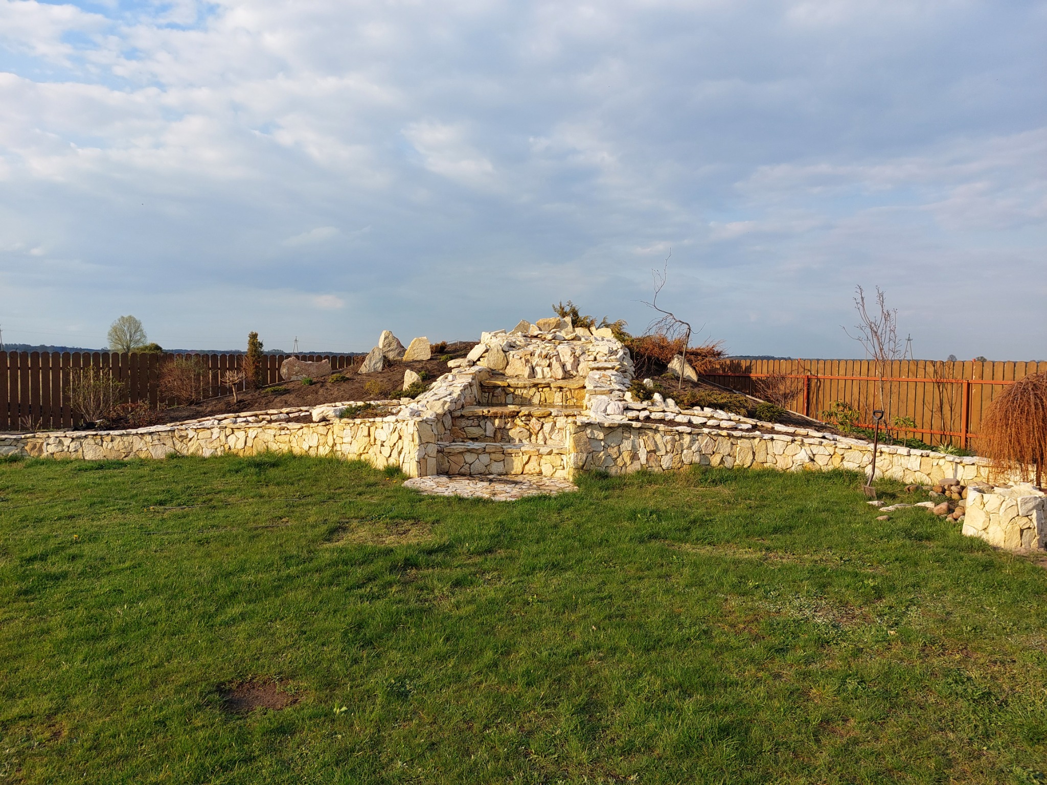 Kaskadowy wodospad ogrodowy z jasnego kamienia na trawniku, otoczony niskim murem oporowym z kamienia, w tle drewniany płot i pochmurne niebo.