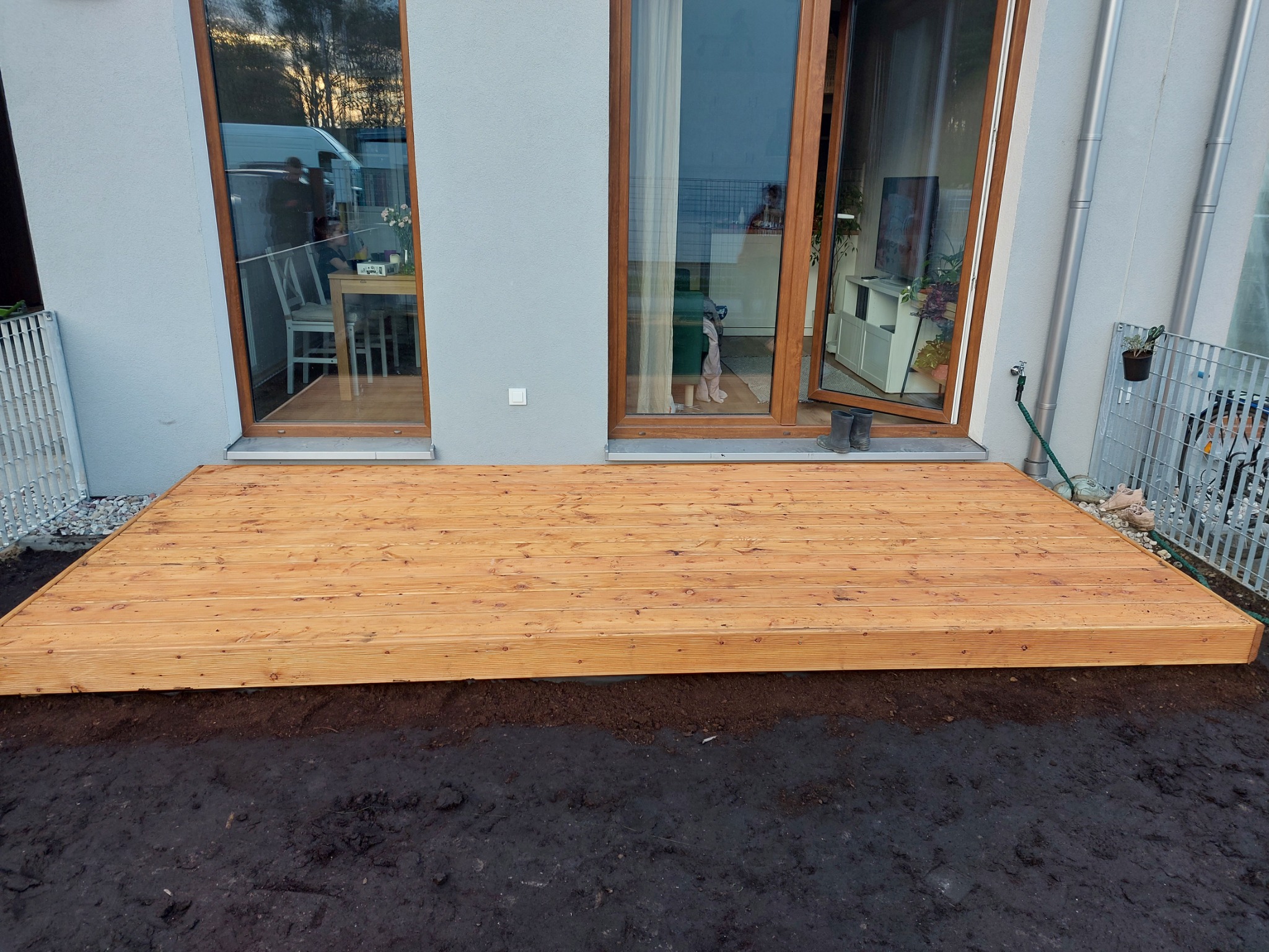 Nowy drewniany taras przydomowy z desek w kolorze jasnego drewna, ziemia wokół, widok na okno tarasowe i wnętrze domu.
