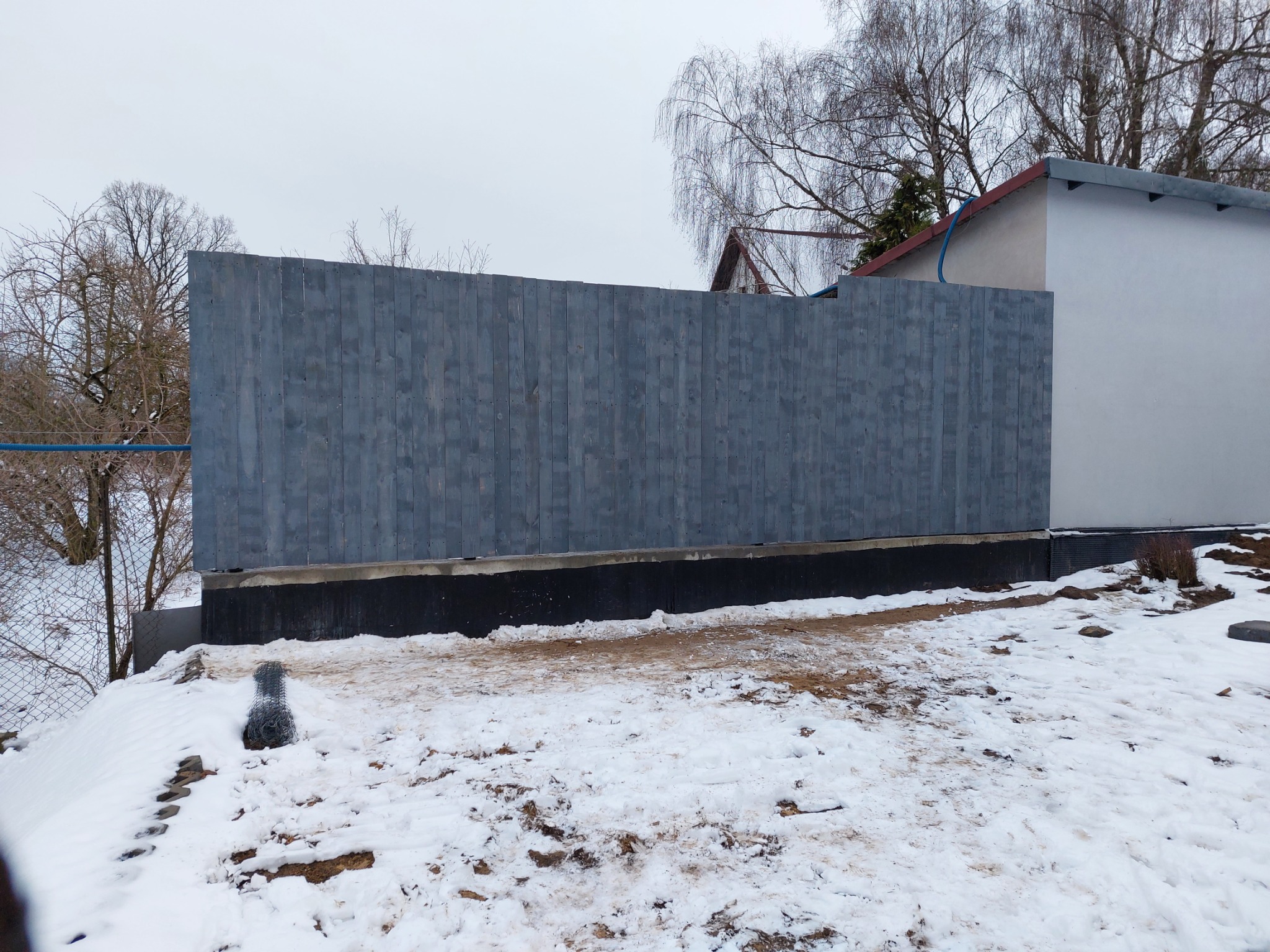 Nowy, pomalowany na szaro drewniany płot panelowy z widocznym fundamentem, otoczony śniegiem i ziemią w zimowy dzień.