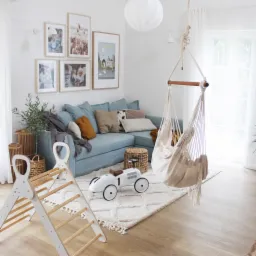 Jasny salon z błękitną sofą narożną, wiszącym hamakiem z frędzlami, drewnianą drabinką gimnastyczną dla dzieci i białym, metalowym samochodem wyścigowym na kremowym dywanie z geometrycznym wzorem.