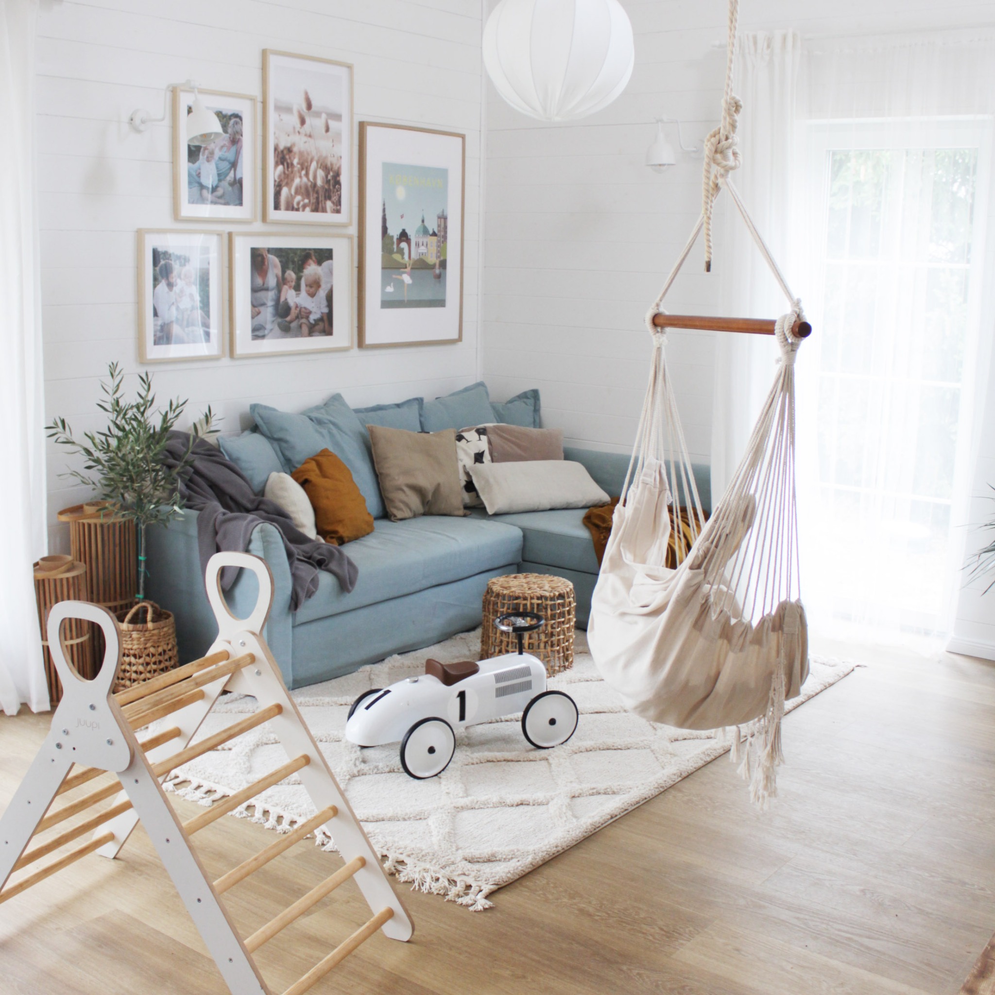 Jasny salon z błękitną sofą narożną, wiszącym hamakiem z frędzlami, drewnianą drabinką gimnastyczną dla dzieci i białym, metalowym samochodem wyścigowym na kremowym dywanie z geometrycznym wzorem.
