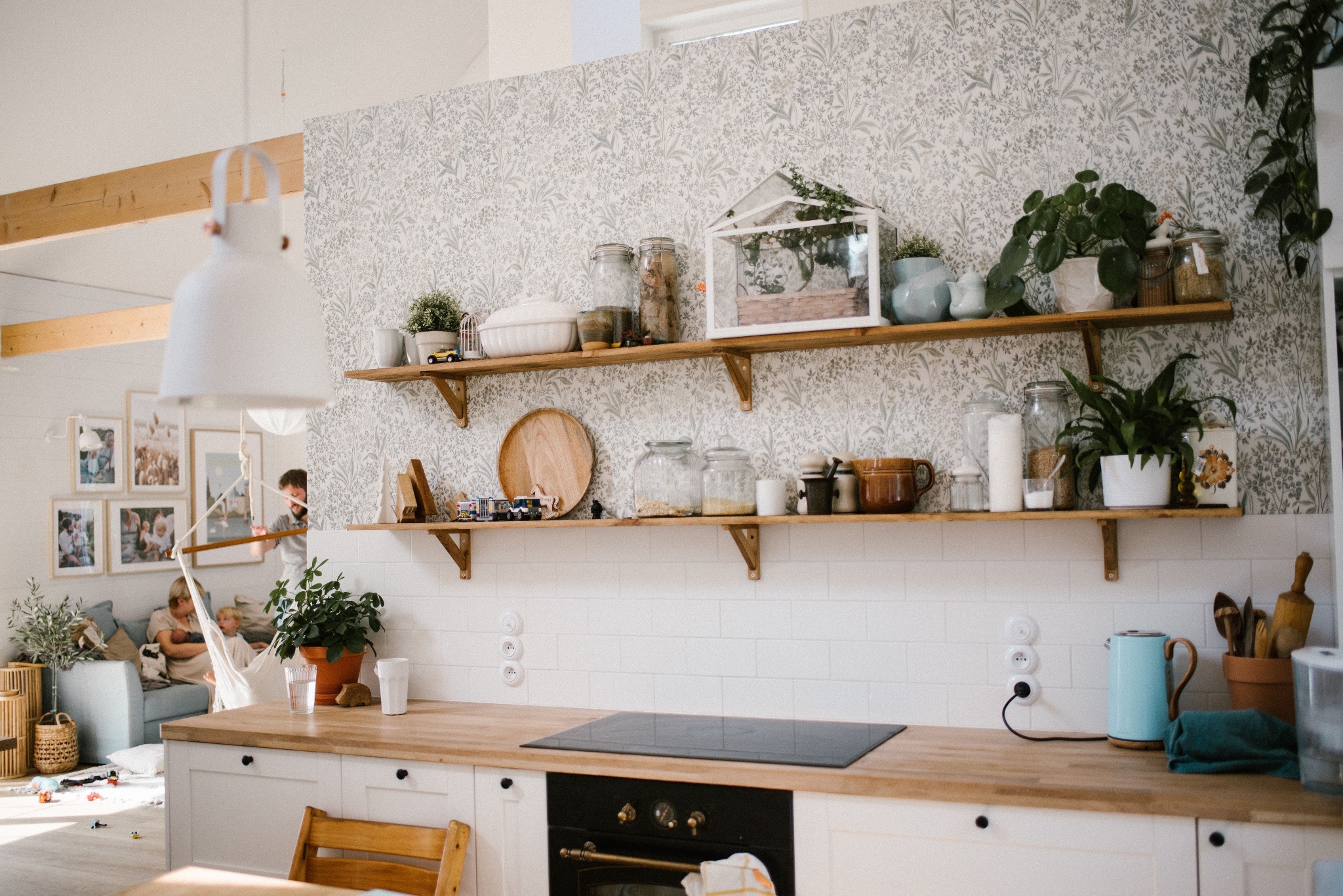 Jasna kuchnia z drewnianym blatem, białymi szafkami i otwartymi półkami z dekoracjami roślinnymi, w tle rodzina w salonie.