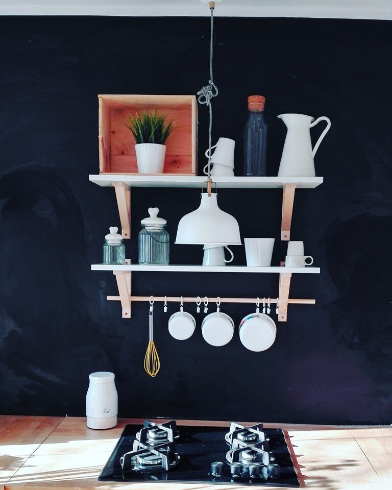 Kuchnia z czarną ścianą tablicową, drewnianymi półkami z białymi naczyniami i rośliną, wiszącymi patelniami i czarną płytą gazową.