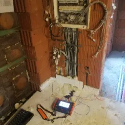 Panel elektryczny w trakcie instalacji w surowym stanie, widoczne przewody, miernik elektryczny z pomarańczowym paskiem i ceglane ściany.