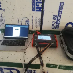 Stanowisko pomiarowe z laptopem Huawei i miernikiem Sonel na tle izolacji Paneltech, widoczna skrzynka transportowa i zawieszone paski do miernika.