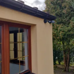 Zewnętrzna kamera monitoringu zamontowana na elewacji domu, widoczna z boku okna z brązowymi ramami i złotymi szprosami, tło stanowi zielone drzewo i metalowa brama.