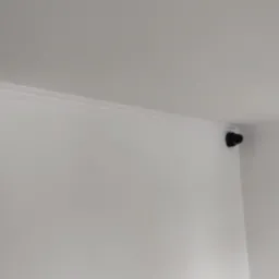 Biały czujnik ruchu i czarna kamera monitoringu zamontowane pod sufitem w rogu jasnego pomieszczenia z oknem.