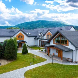 Rozległy, nowoczesny dom z szarą dachówką i jasną elewacją, otoczony zielonym trawnikiem, krzewami i drzewami iglastymi, z górą w tle pod błękitnym niebem z chmurami.