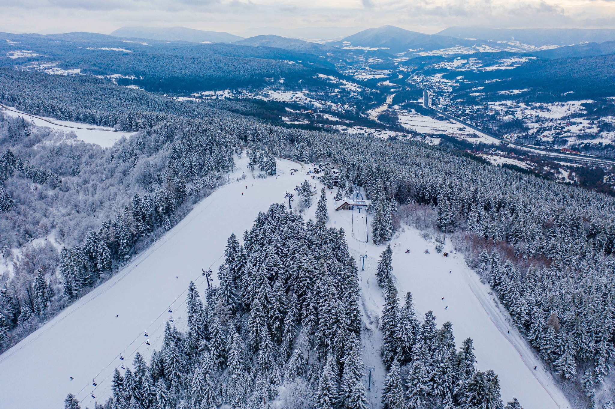 Widok z lotu ptaka na zasypany śniegiem stok narciarski z wyciągami krzesełkowymi, otoczony lasem iglastym i górami w tle, pod pochmurnym niebem.