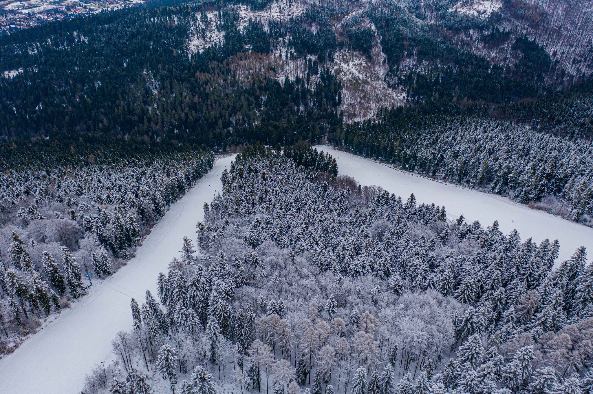 Zimowy krajobraz z lotu ptaka: ośnieżone lasy iglaste i polany pokryte śniegiem, widoczny wyciąg narciarski i niewielkie zabudowania w oddali.