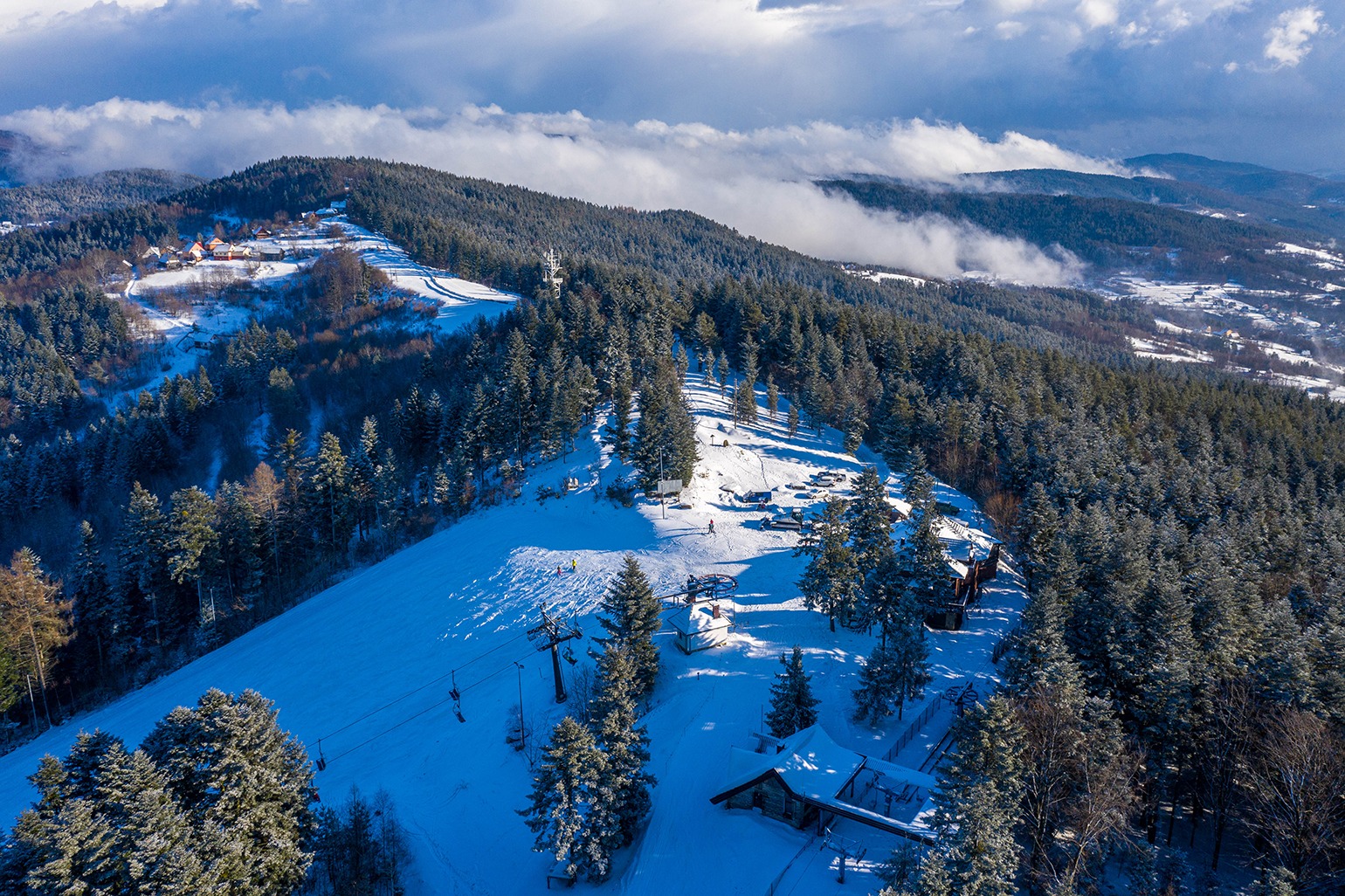 Zimowy krajobraz górski z widokiem z lotu ptaka na zaśnieżony stok narciarski, las iglasty i wyciąg krzesełkowy w słoneczny dzień.