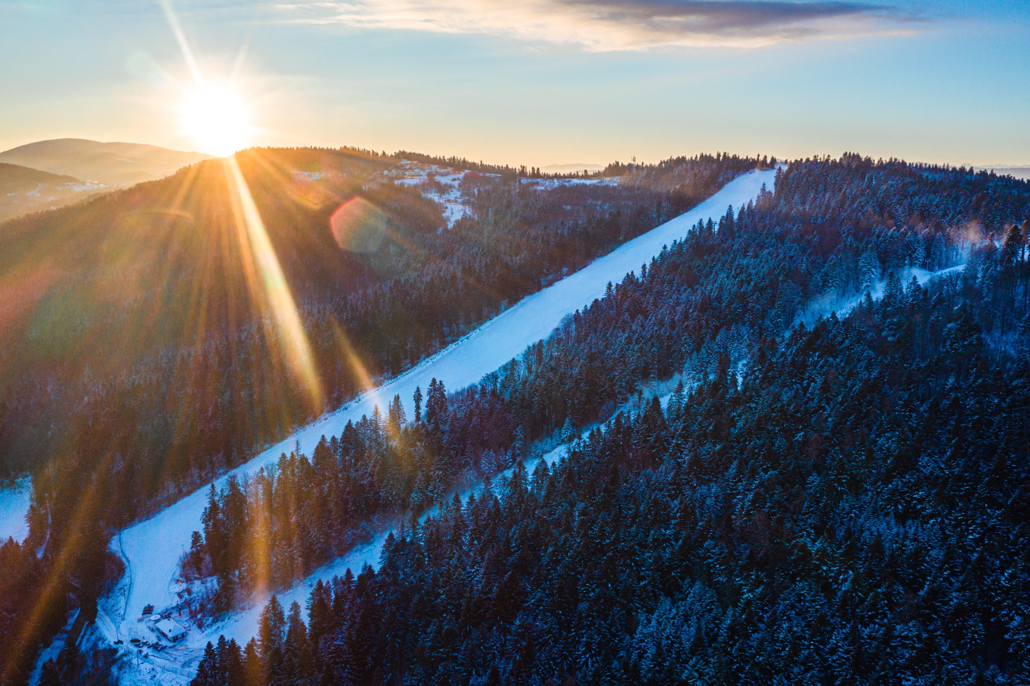 Widok z lotu ptaka na ośnieżony stok narciarski w otoczeniu lasu iglastego, oświetlony promieniami zachodzącego słońca, z widocznymi budynkami u podnóża stoku.