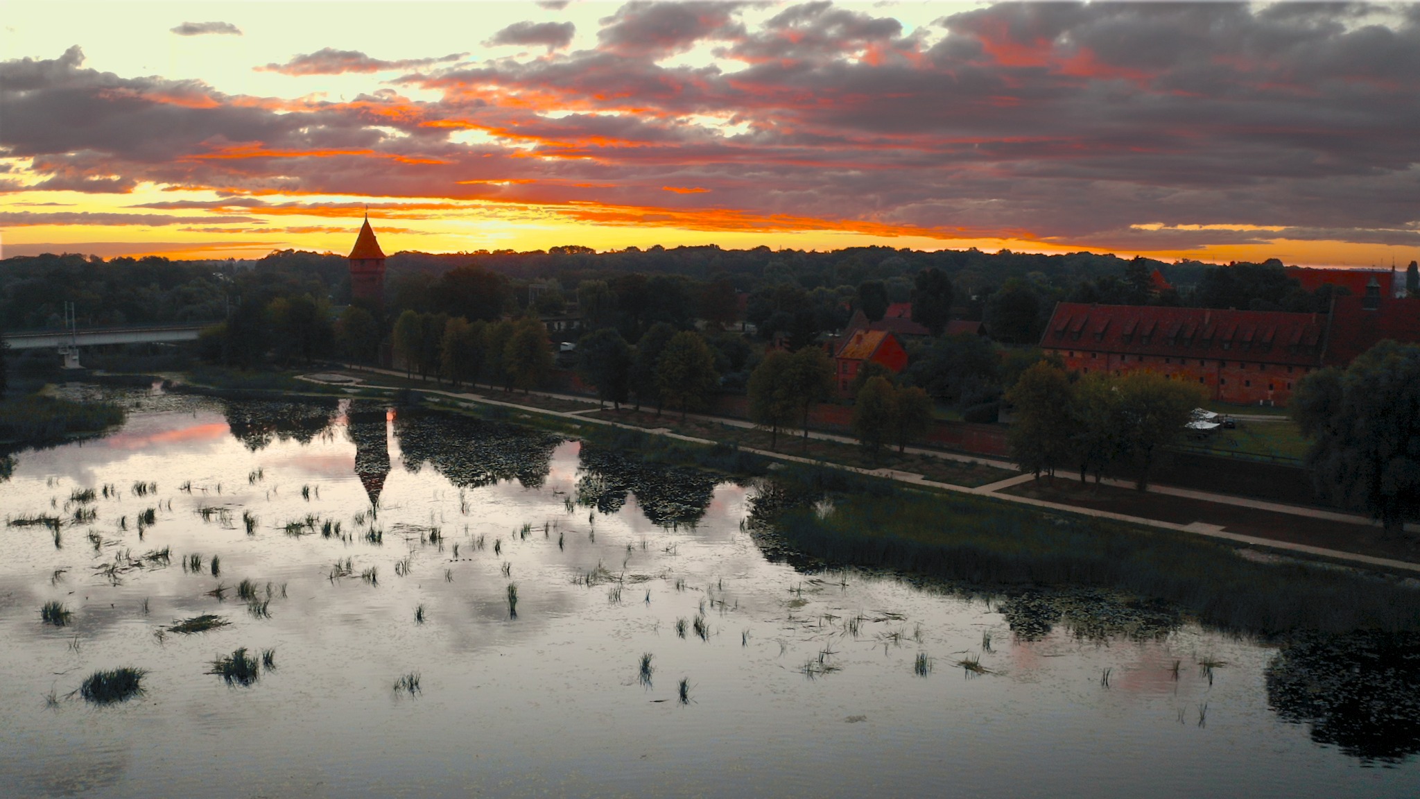Panorama zamku nad rzeką o zachodzie słońca, z wieżą odbijającą się w wodzie i dramatycznym, pomarańczowo-szarym niebem w tle.