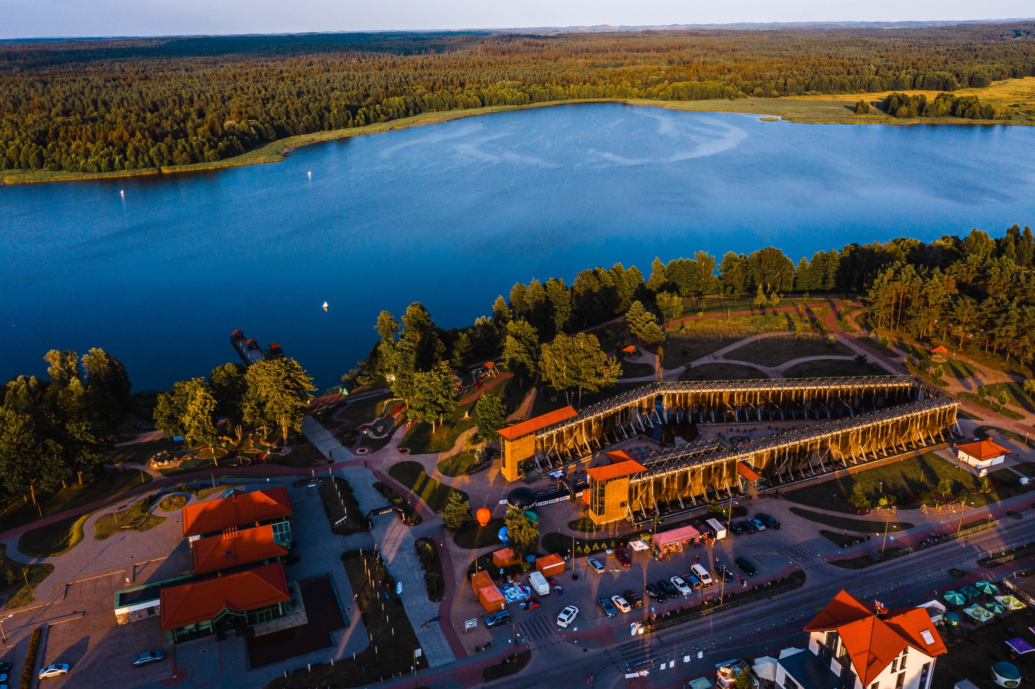 Panoramiczne ujęcie z lotu ptaka na jezioro z lasem w tle i kompleks solankowy z drewnianą konstrukcją oraz budynkami z czerwonymi dachami, oświetlone ciepłym światłem zachodzącego słońca.