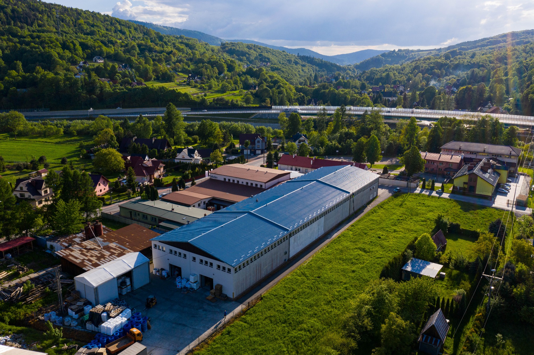 Widok z lotu ptaka na budynek magazynowy z niebieskim dachem, otoczony zielenią, z widoczną autostradą i górami w tle.