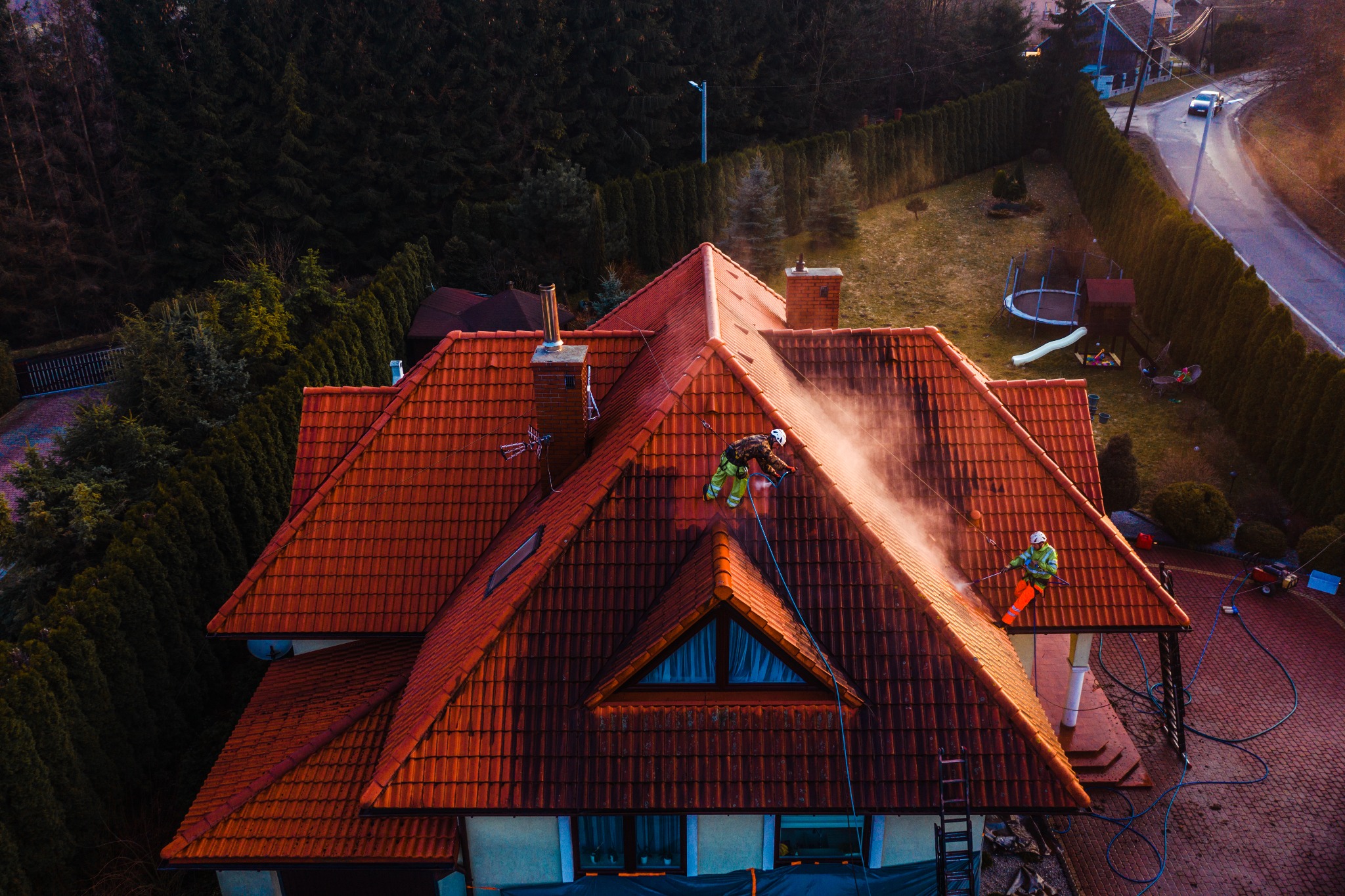 Dwóch pracowników w uprzężach czyści dach z czerwonej dachówki za pomocą myjek ciśnieniowych, widok z lotu ptaka na dom z ogrodem.